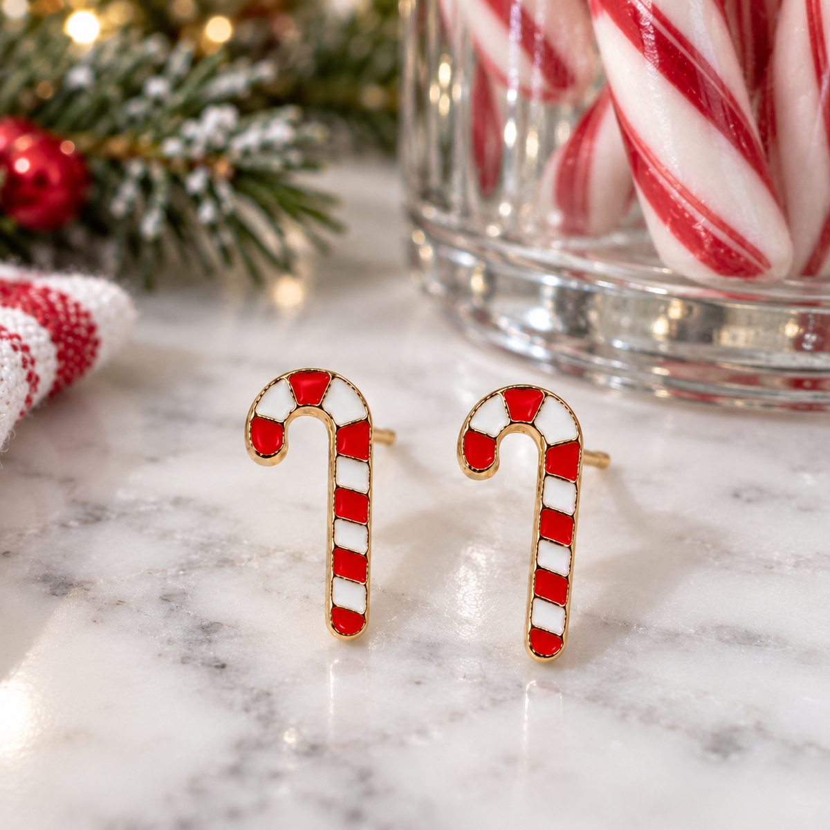 A pair of gold-tone Classic Striped Candy Cane Earrings with red and white striped enamel, displayed on a white marble surface with holiday decorations and a glass jar of candy canes in the background.