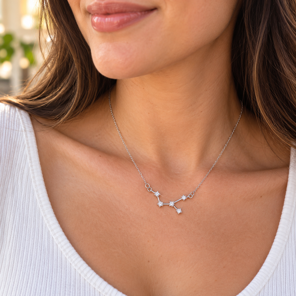 A close-up of a woman wearing a delicate silver constellation zodiac necklace with a small pendant featuring tiny clear stones, styled with a white ribbed top.