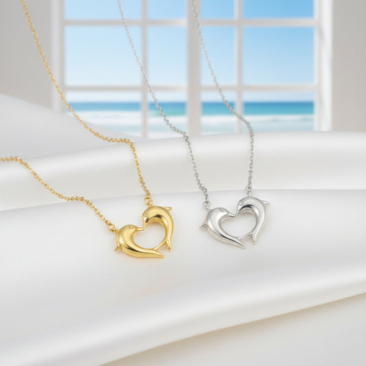 Gold and silver Dolphin Heart Necklaces on a light surface with a window in the background.
