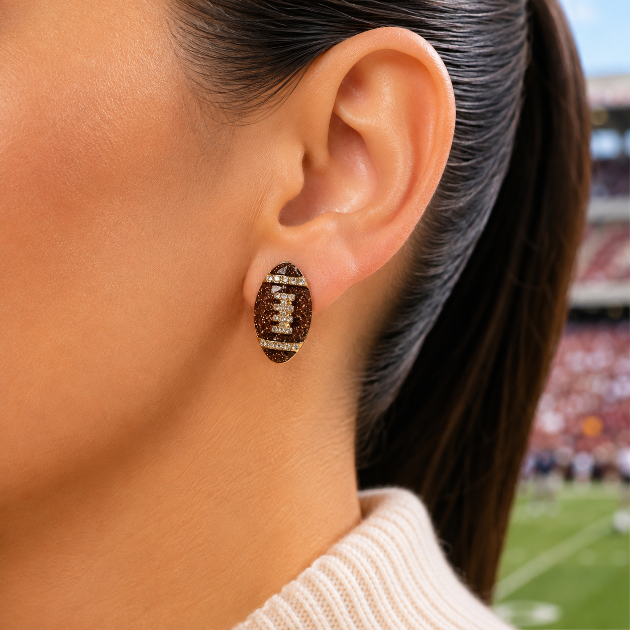 A close-up of a woman's ear wearing a Game Day Sparkle Football Earring, set against a blurred stadium background.