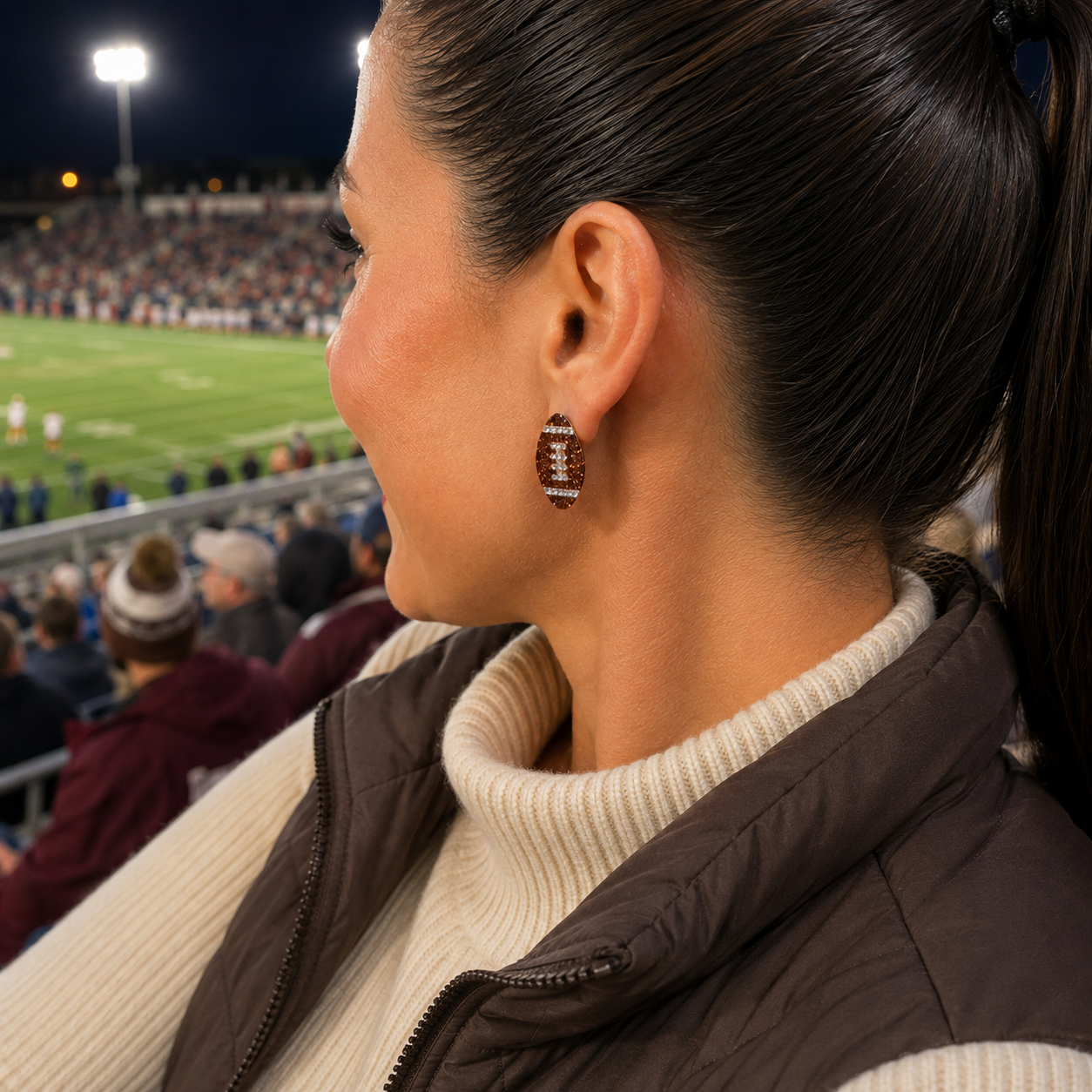 A woman in a stadium wearing Game Day Sparkle Football Earrings, shown in side profile with a football field and crowd in the background at night.