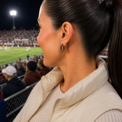 A close-up side profile of a woman at a night football game, wearing a Game Day Sparkle Football Earrings and a cream puffer vest.