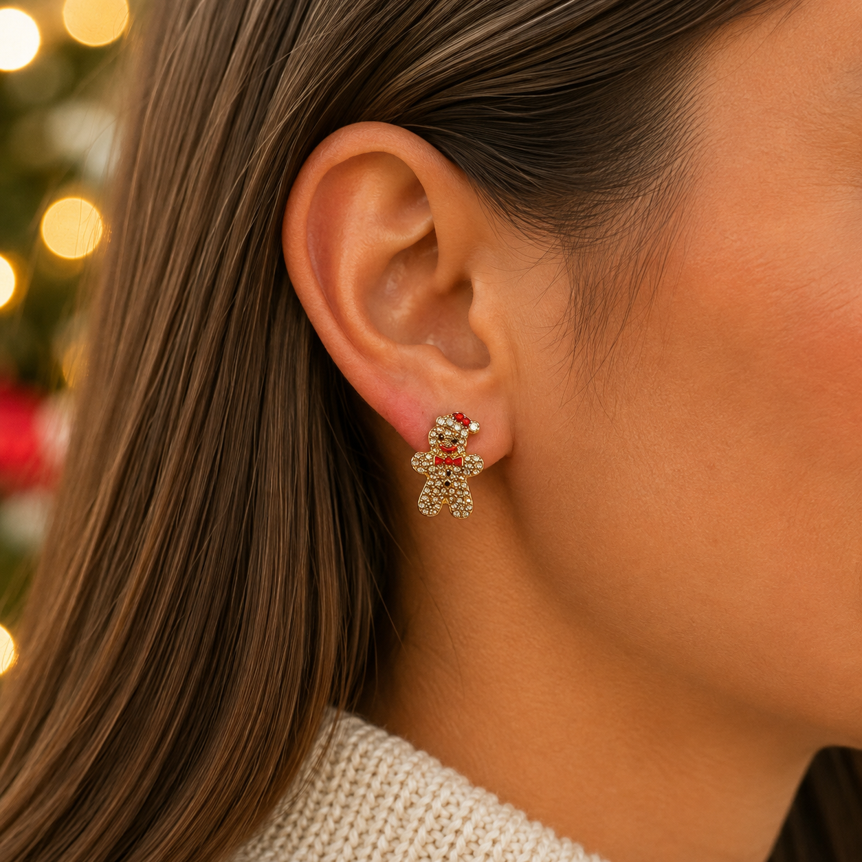 A close-up of a woman's ear wearing a Gingerbread Man Pavé Earring, with tiny clear stones and a red stripe detail, against a softly blurred holiday background.