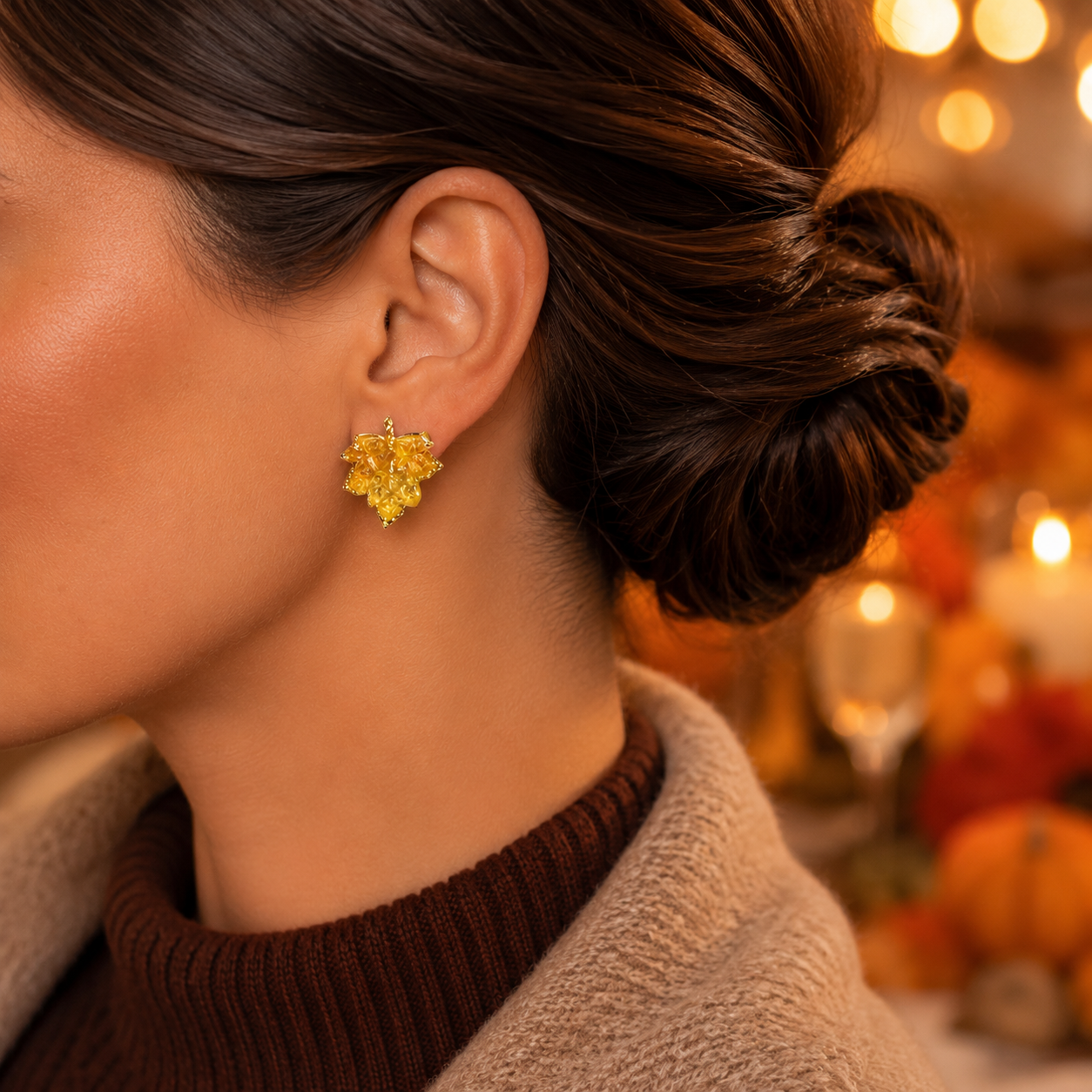 A close-up side profile of a woman wearing a Golden Maple Leaf Earring, with her hair in a bun and a cozy brown sweater visible against a warm, softly blurred background with lights and pumpkins.