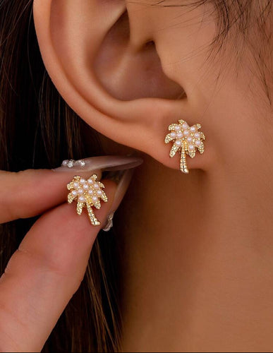 A pair of gold-tone palm tree earrings accented with white imitation pearls, worn on a person's ear.