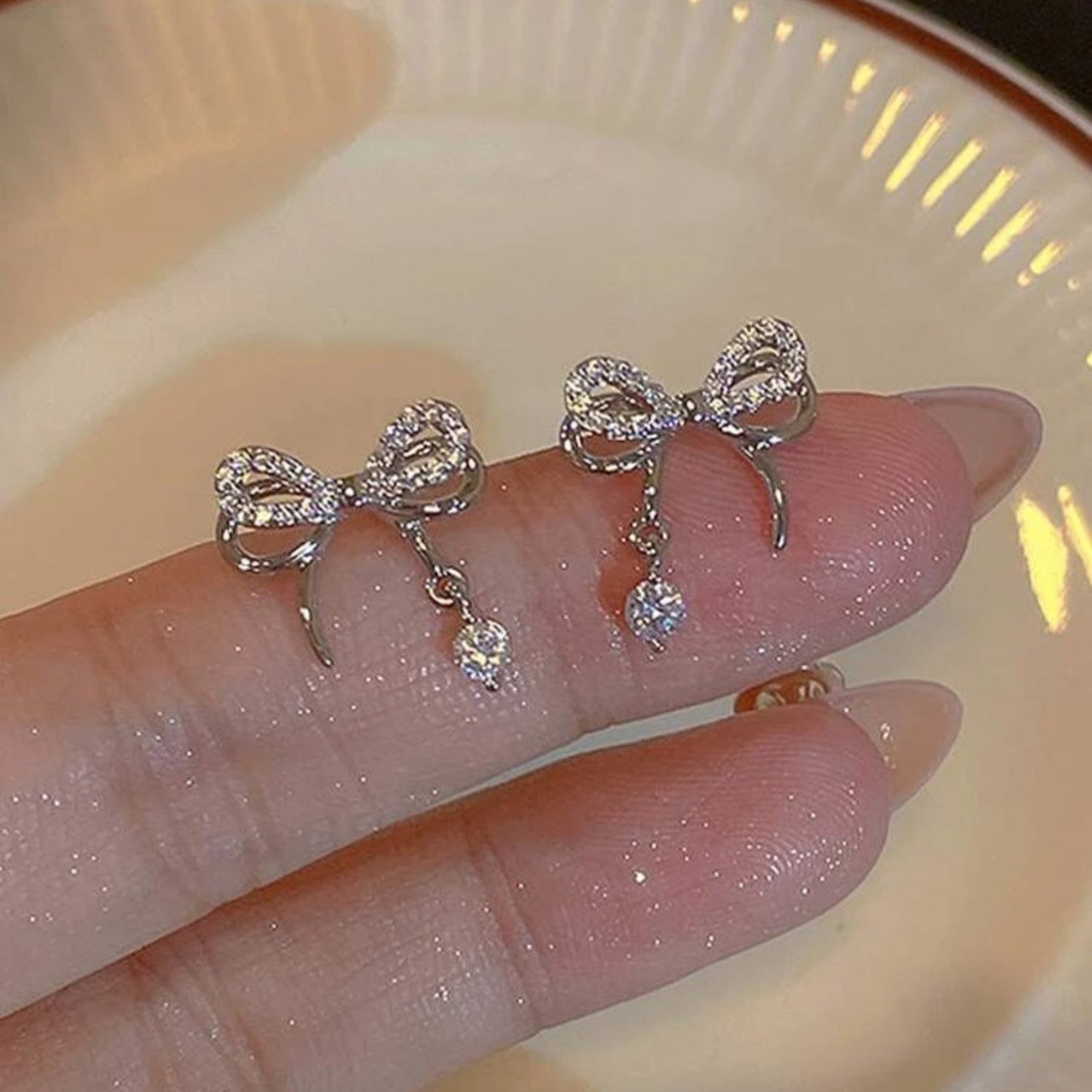 Silver bow-shaped earrings with dangling crystals on a hand against a neutral background
