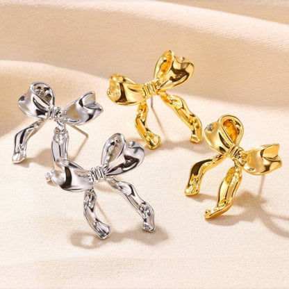 Two pairs of Silver Bow Ribbon Earrings, one gold and one silver, on a beige background.