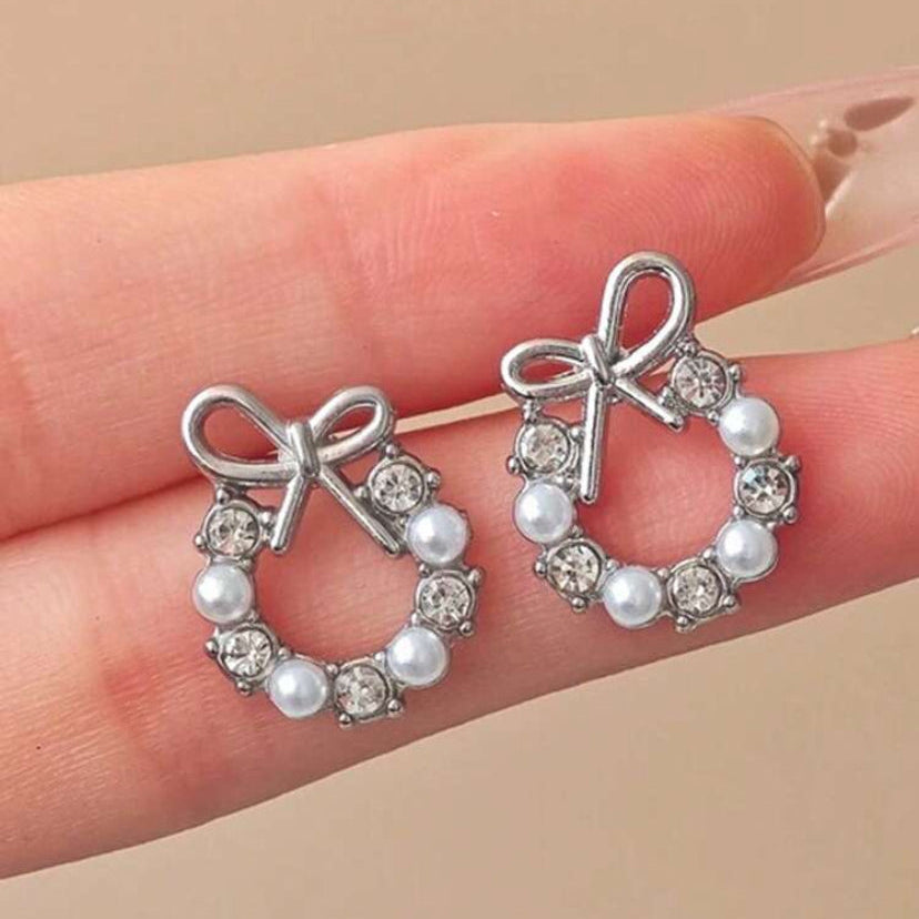 Silver Bow Wreath Earrings with bow and pearl design held between fingers on a beige background