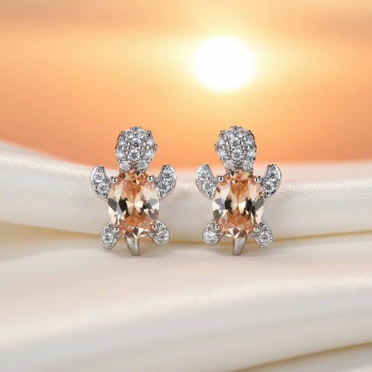 Orange Glow Crystal Turtle Earrings with orange gemstones and silver accents on a fabric and sunset background