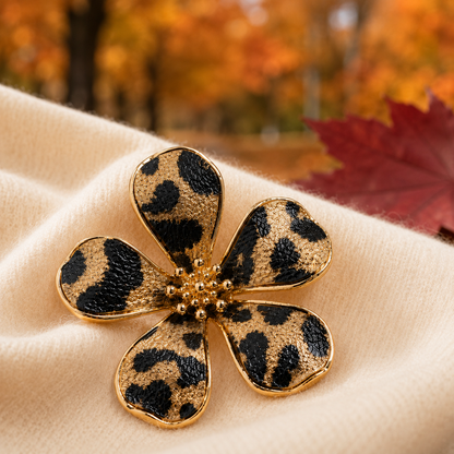 A Leopard Print Flower Pin with a leopard print pattern and gold-tone metal borders, resting on a soft beige fabric with a blurred autumn leaf background.