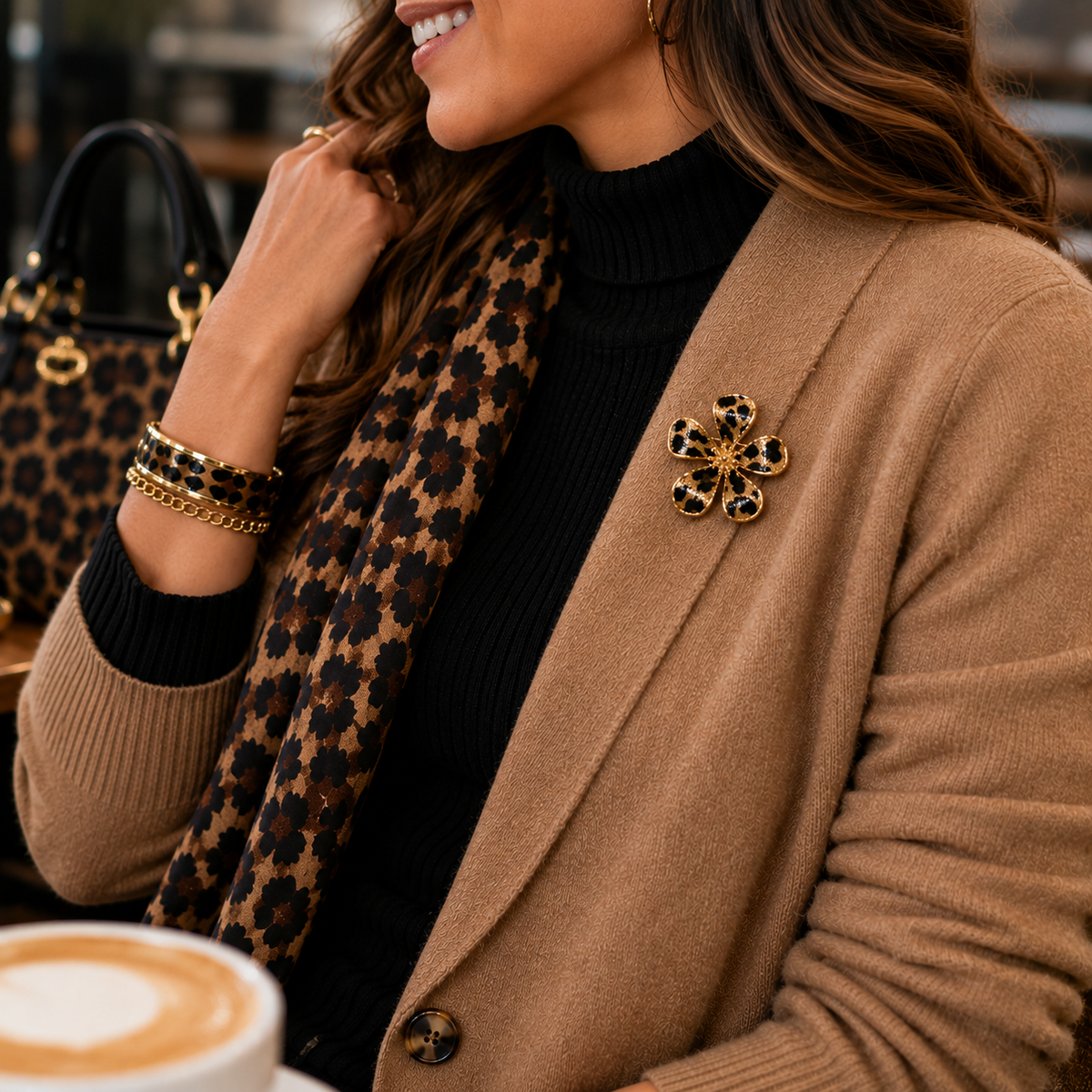A woman in a cafe setting wearing a tan wool-blend cardigan, a black turtleneck, a leopard-print scarf, and a Leopard Print Flower Pin. A matching leopard-print handbag is visible in the background.