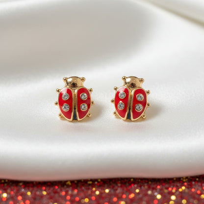 Gold and red Little Lucky Ladybug Earrings on a white surface with a red glittery background.