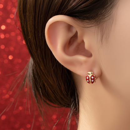 Close-up of an ear wearing a red and gold Little Lucky Ladybug Earrings against a red glittery background