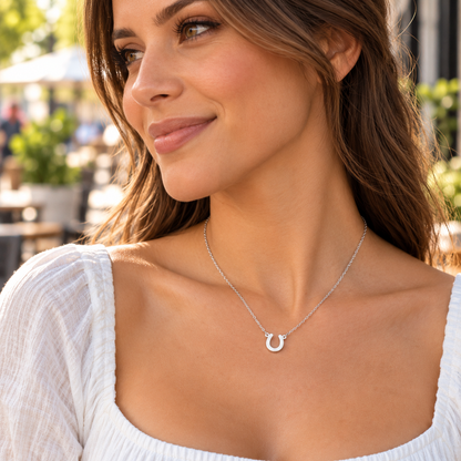 A woman wearing a Lucky Horseshoe Necklace, shown in a close-up profile view. She is wearing a white textured top, and the background is a softly blurred outdoor patio setting.