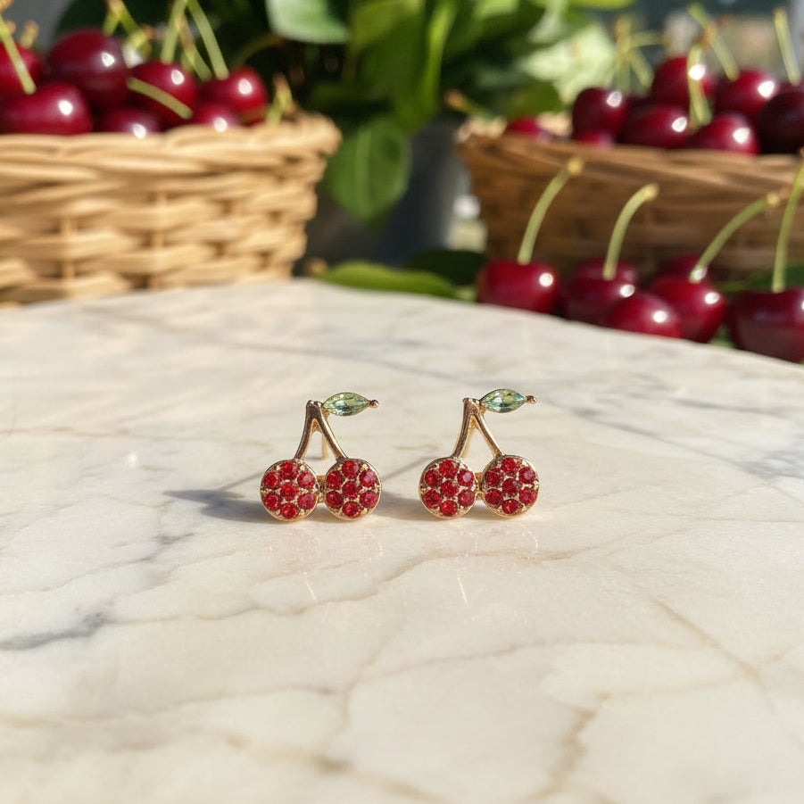 Mini Red Pavé Cherry Earrings on a marble surface with baskets of cherries in the background.