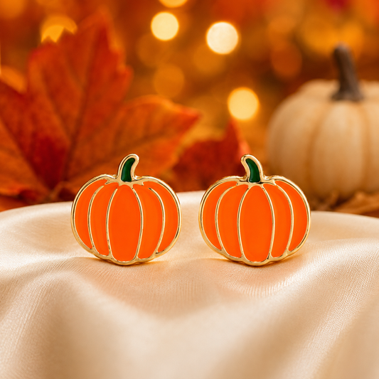 A pair of Orange & Gold Pumpkin Earrings, displayed on cream satin fabric with autumn leaves and a blurred pumpkin in the background.