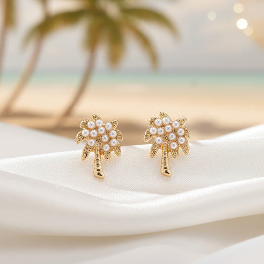 A pair of gold-tone palm tree earrings accented with white imitation pearls, displayed on a white textured surface.