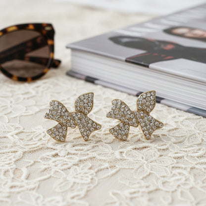 Pavé Crystal Bow Earrings on a textured surface with sunglasses and book in the background.