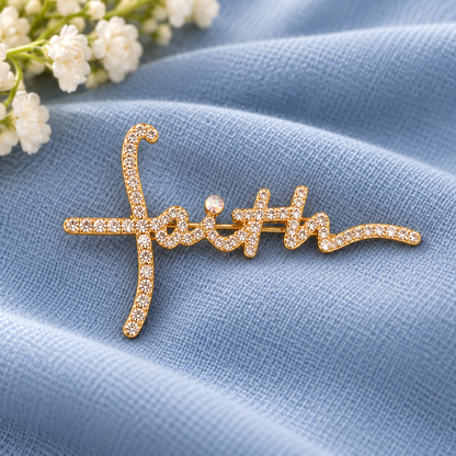 A gold cursive word-art brooch spelling 'faith' with pavé-set clear stones, displayed on a textured light blue fabric with small white flowers in the corner.