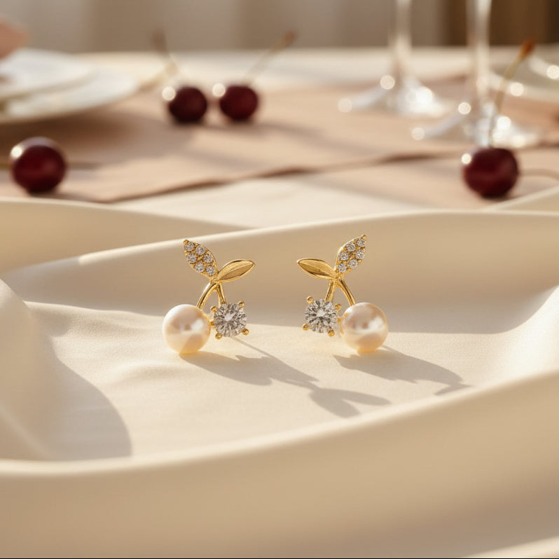 A pair of gold-tone Pearl Cherry Earrings with zircon accents on a fabric surface.
