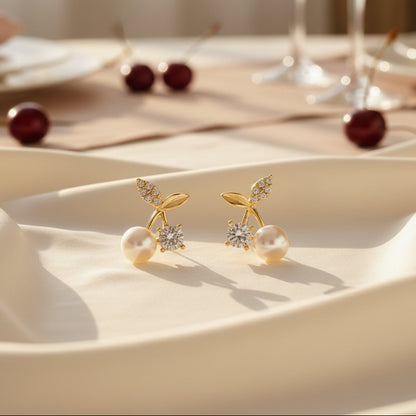 A pair of gold-tone Pearl Cherry Earrings with zircon accents on a fabric surface.