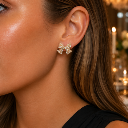 A close-up of a woman's ear wearing a Pearl & Pavé Bow Earrings, against a softly blurred warm indoor background.