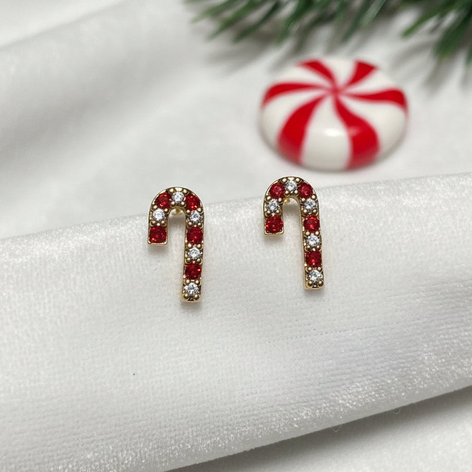 A pair of candy cane-shaped stud earrings with red stones and clear rhinestones in a gold-tone setting, displayed on a white card held against a person's fingers.