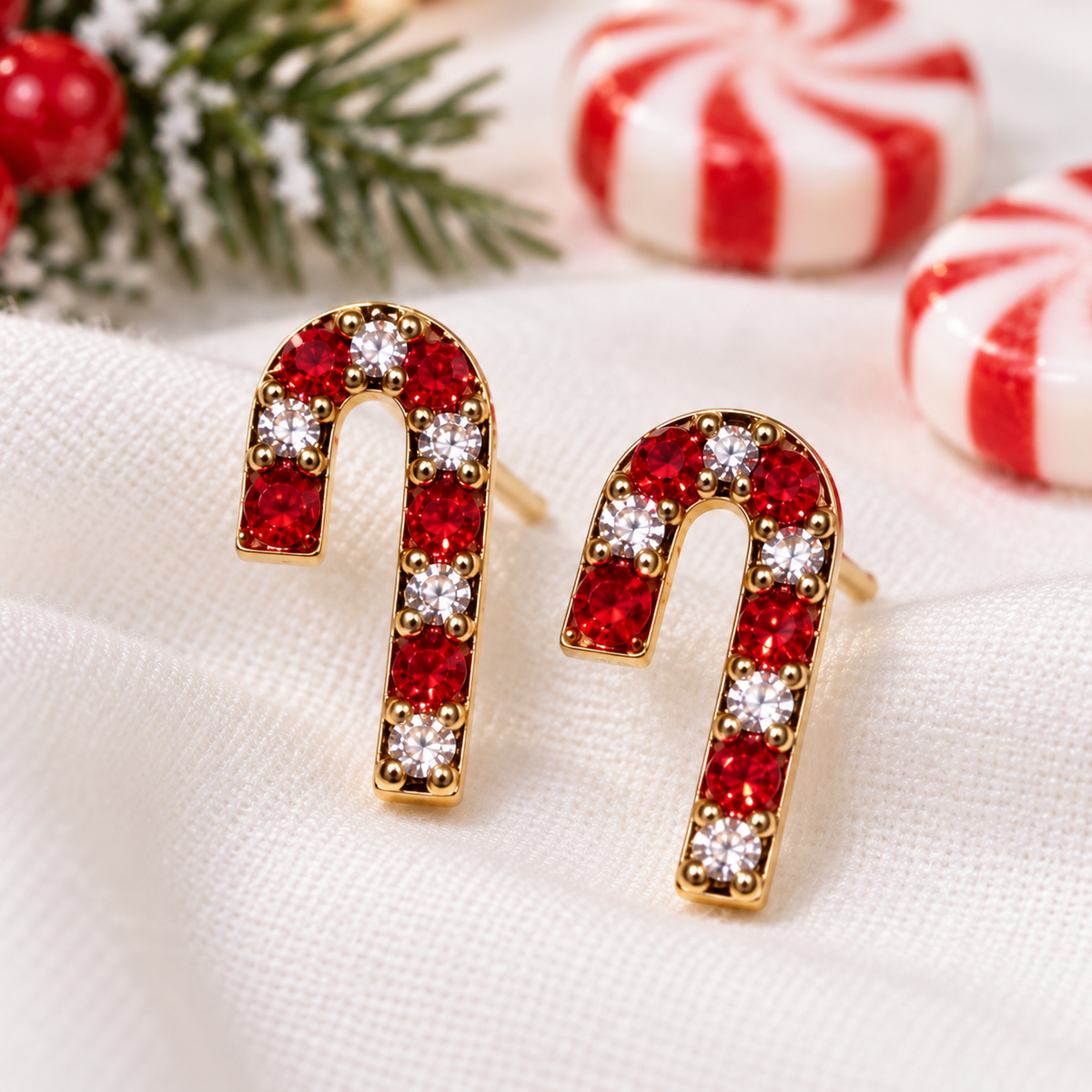 Gold Peppermint Pavé Candy Cane Earrings with red and clear rhinestones, displayed on a white textured fabric with festive holiday decorations in the background.