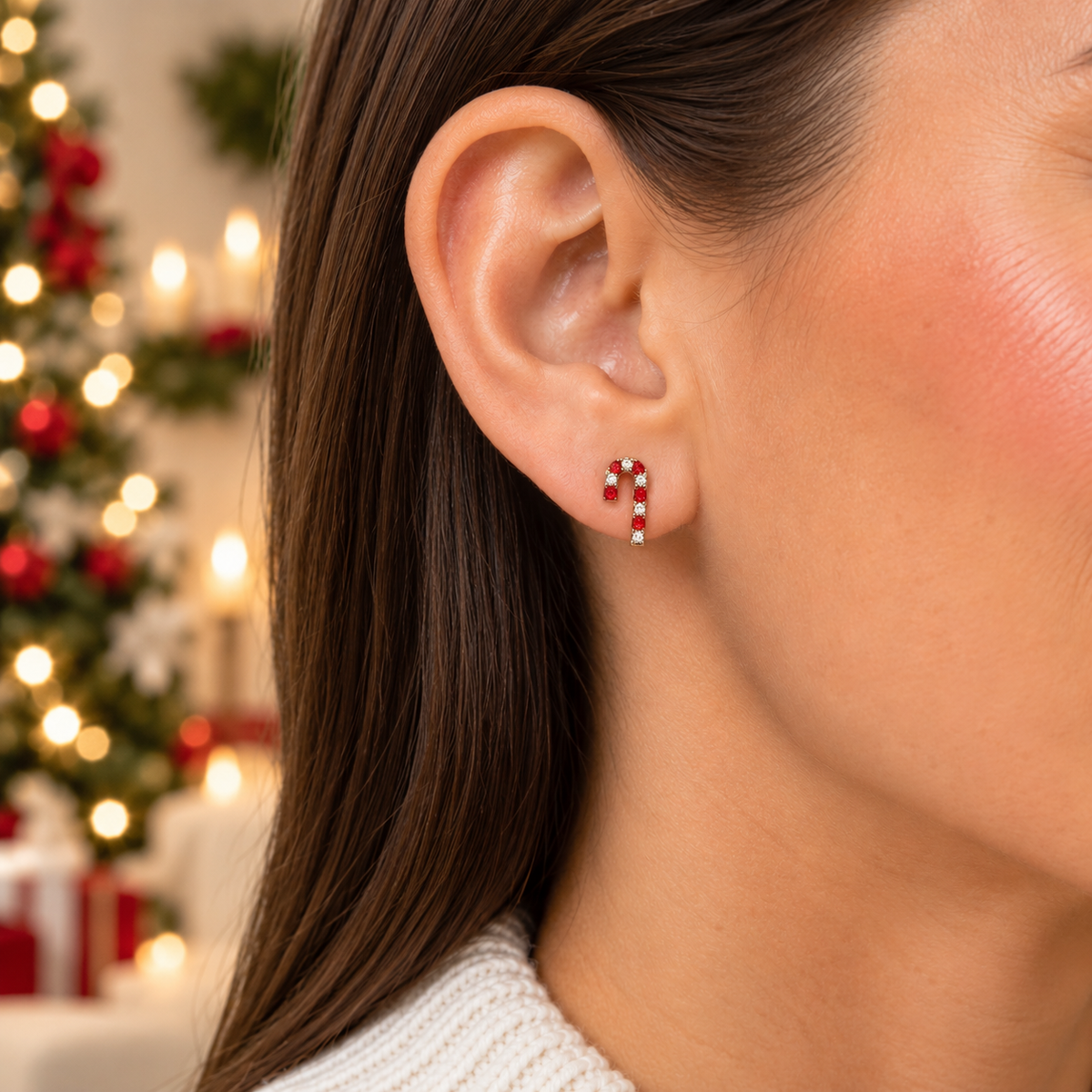 A close-up of a woman's ear wearing a small Peppermint Pavé Candy Cane Earring with red and white stones, set against a softly blurred Christmas tree background.