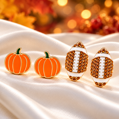 Pumpkin & Football Pavé Earrings Bundle displayed on draped white satin fabric, with a warm blurred autumn background.
