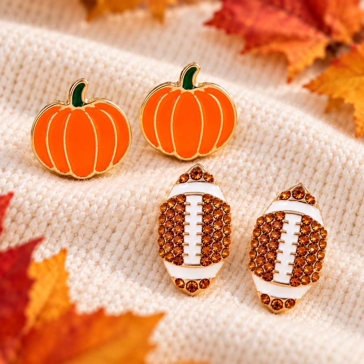 Pumpkin & Football Pavé Earrings Bundle arranged on a cream knit fabric with autumn leaves. The pumpkin earrings are orange with green stems and gold-tone outlines, while the football earrings are white with gold-tone outlines and covered in orange rhinestones.