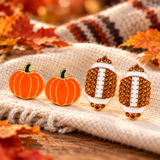 Pumpkin & Football Pavé Earrings Bundle on a cream woven textile with autumn leaves around them: two pumpkin-shaped earrings with orange enamel and green stems, and two football-shaped earrings covered in amber rhinestones with white center detailing.