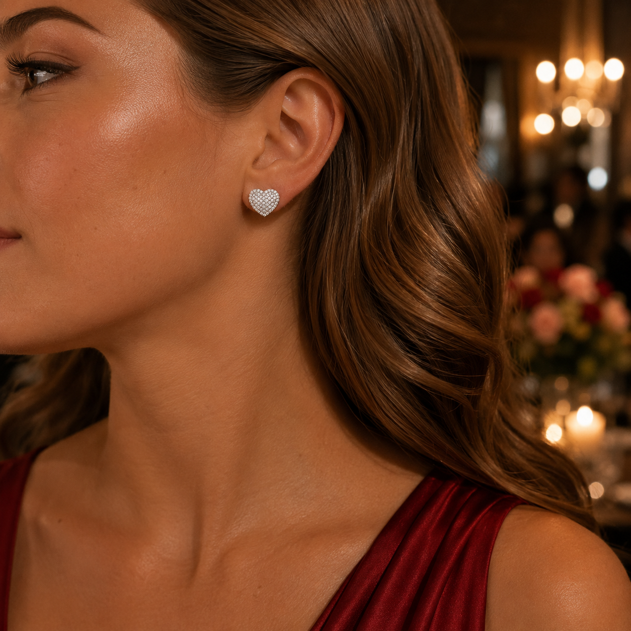 A close-up of a woman wearing a Radiant Heart Pavé Earring. The earring is shown on her earlobe in a warm, elegant indoor setting with blurred lights in the background.