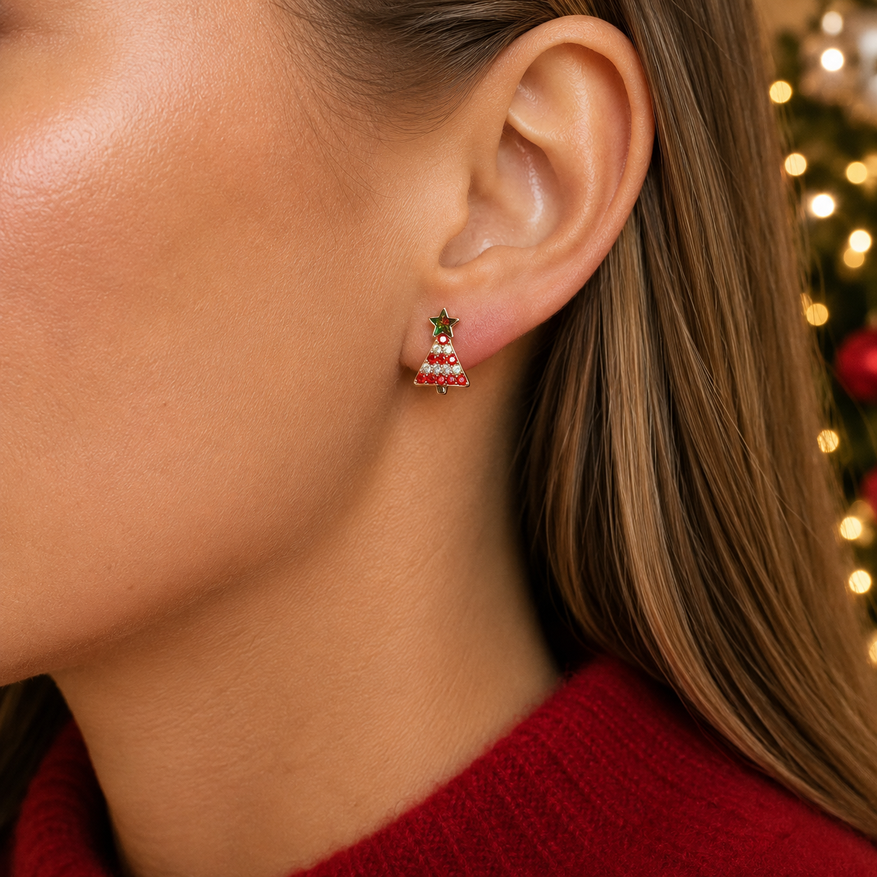 A close-up of a woman's ear wearing a Red Pavé Christmas Tree Earring with red and white stripes and a green star on top, against a softly blurred holiday background with warm lights.