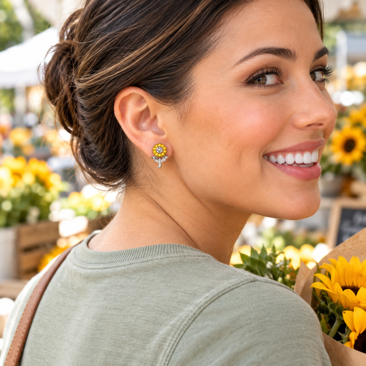 A woman with her hair in a bun is shown in profile at a flower market, wearing a Sunflower Sparkle Earring. She is wearing an olive green shirt and holding a bouquet of sunflowers.