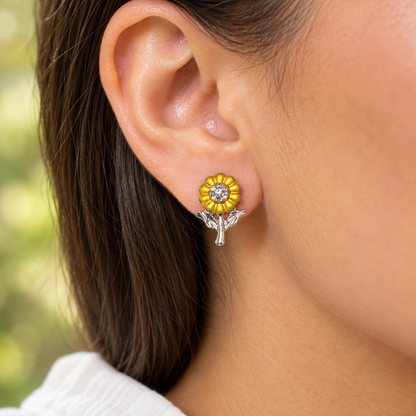 A close-up of a person's ear wearing a Sunflower Sparkle Earrings with yellow petals, a clear center stone, and silver-toned leaves.
