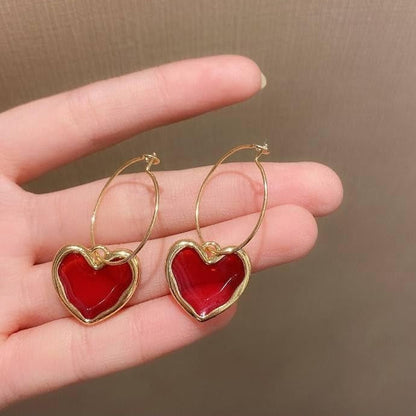 Ruby Heart Hoop Earrings with red centers and gold frames held in a hand against a brown background