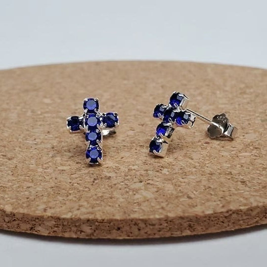 Silver Mini Blue Crystal Cross Earrings with blue gemstones on a cork board.