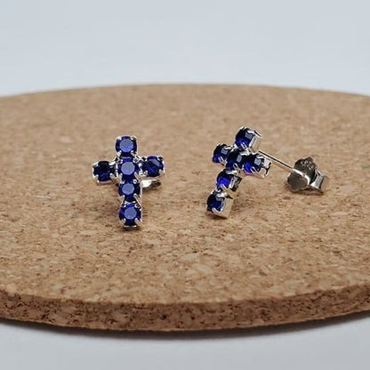 Silver Mini Blue Crystal Cross Earrings with blue gemstones on a cork board.