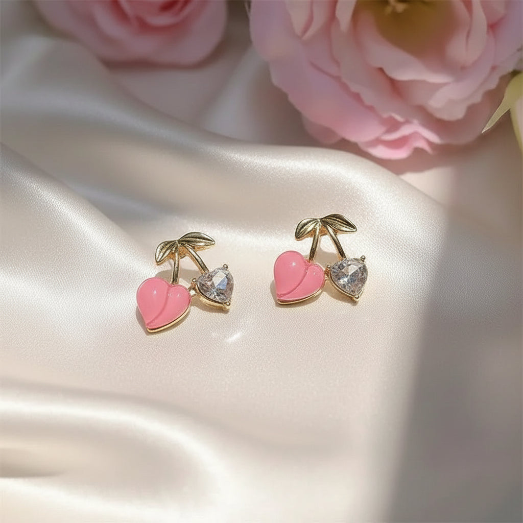 Pink Cherry & Crystal Heart Earrings with pink and gold details on a light surface with flowers in the background